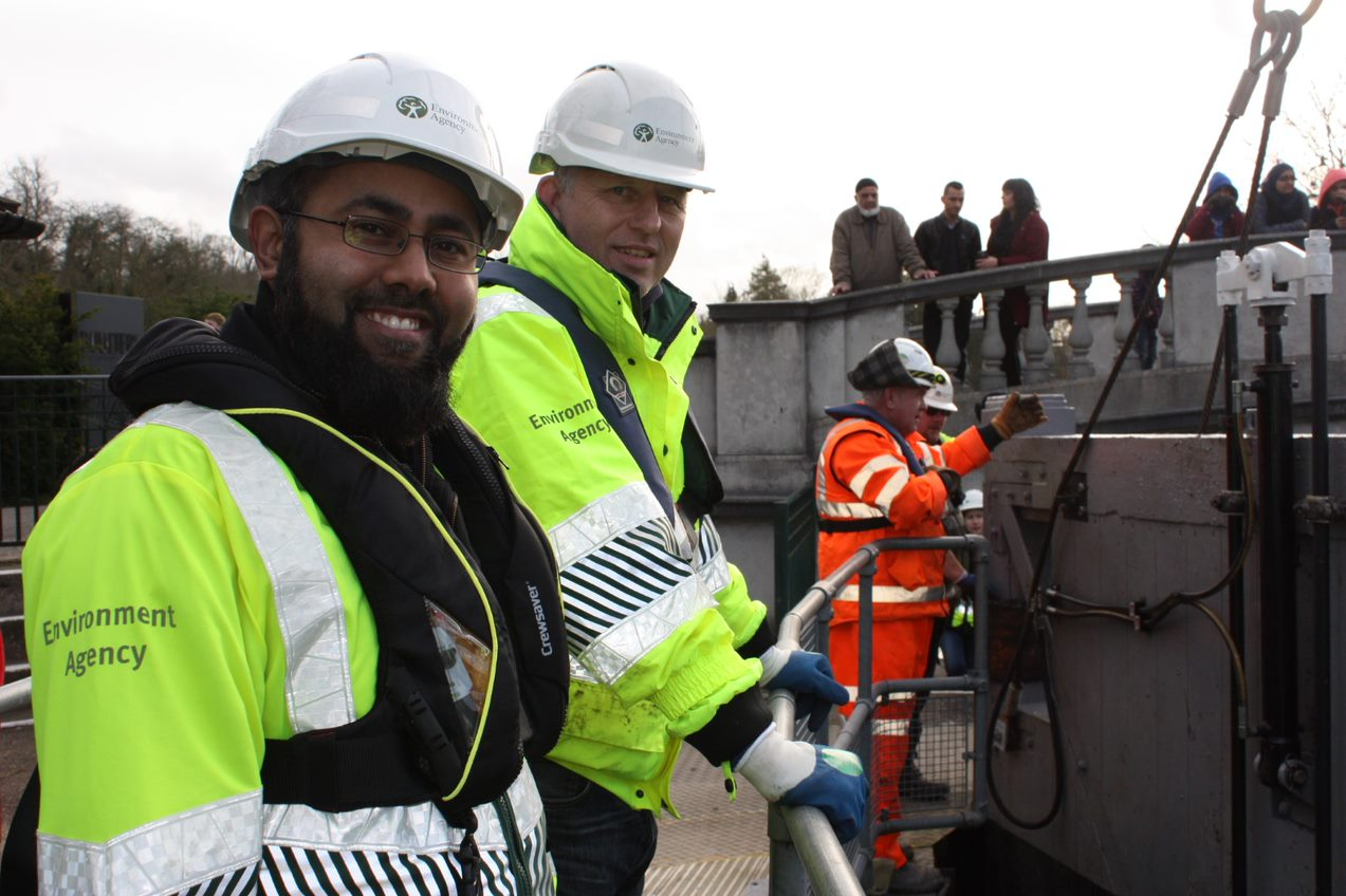 Two Environment Agency Staff facing right while looking at the camera in yellow branded hi-vis. In the background are three workers in orange hi-vis assessing their work, under and beside a bridge full of onlookers.